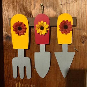 May include: Three wooden garden tools with yellow, red, and gray paint. Each tool has a sunflower-shaped flower on top. The tools are a fork, a trowel, and a hand rake.