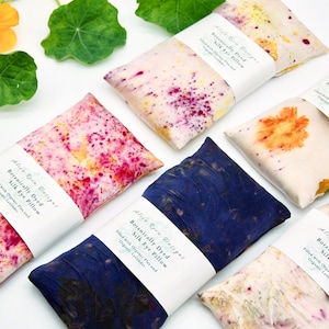 May include: A selection of silk eye pillows, each dyed with botanical patterns in pink, blue, and yellow hues. The rectangular pillows are labelled with "Botanically Dyed Silk Eye Pillow".
