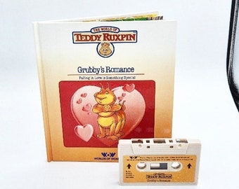 May include: A vintage children's book titled "Grubby's Romance" by Teddy Ruxpin, with a matching cassette tape. The book cover features an illustration of a Grubby character with hearts. The cassette tape has the same title.