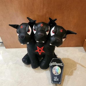 May include: A black three-headed plush dog with red pentagram embroidery on the chest and red number sixes on each forehead. The toy features white fangs and a coffin-shaped tag labeled Killstar Kreepures with text including 1982 and I'm cute as hell.