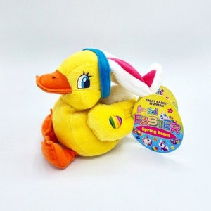 Puede incluir: Un pato de peluche amarillo con una diadema azul y orejas de conejo con detalles en rosa y blanco. El pato tiene pico y patas naranjas, y un corazón arcoíris en su ala. Lleva una etiqueta de Lisa Frank Easter.