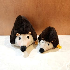 May include: Two plush hedgehog toys. The hedgehogs have dark brown fur on their backs, cream-colored faces, and black noses. They have small, round eyes and tiny ears. One is larger than the other. Both have yellow tags.