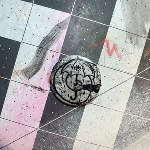 May include: A black and white button with a graphic of an umbrella with an eye and a hand holding it. The umbrella is over a heart.