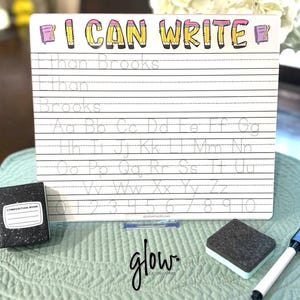 May include: A white rectangular dry-erase board with the text I CAN WRITE in yellow and pink pencil-themed letters. The board features black lined practice rows with dotted letters from A to Z and numbers 0 to 10, a small black composition book, a felt eraser, and a marker.