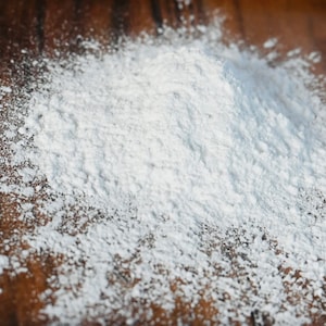 May include: A close-up of white powder, such as flour or icing sugar, scattered on a brown wooden surface.
