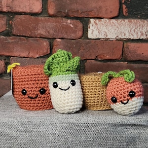 May include: Four crocheted vegetable-shaped items with smiling faces. The vegetables include a carrot, a radish, a turnip, and a tomato. The items are in shades of orange, white, and green. The background is a brick wall.