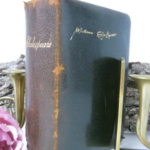 Op de afbeelding: Een vintage boek getiteld "Shakespeare" met een donkergroene kaft en gouden letters. Het boek wordt ondersteund door een messing boekensteun. De rug van het boek is bruin met zichtbare slijtage, wat duidt op de leeftijd.