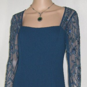 May include: A teal formal gown with a square neckline and sheer sleeves embellished with blue beading. The floor-length dress is accessorized with a silver necklace and green pendant. The dress is displayed on a mannequin.