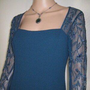 May include: A teal formal dress with a square neckline and sheer sleeves. The sleeves feature a floral beaded design. A silver necklace with a green pendant complements the dress.