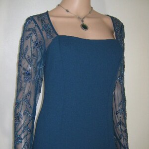 May include: A teal formal dress with a square neckline and long, sheer sleeves. The sleeves feature detailed beadwork. A silver necklace with a pendant is also visible.