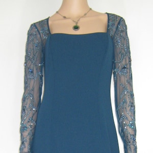May include: A teal evening gown with a square neckline and long sleeves. The sheer sleeves are embellished with detailed beadwork. A delicate necklace with a pendant complements the dress. The dress has a floor-length skirt.