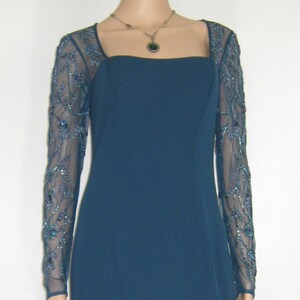 May include: A teal evening gown with a square neckline and long, sheer sleeves adorned with blue beading. The dress features a floor-length skirt and is shown on a mannequin.