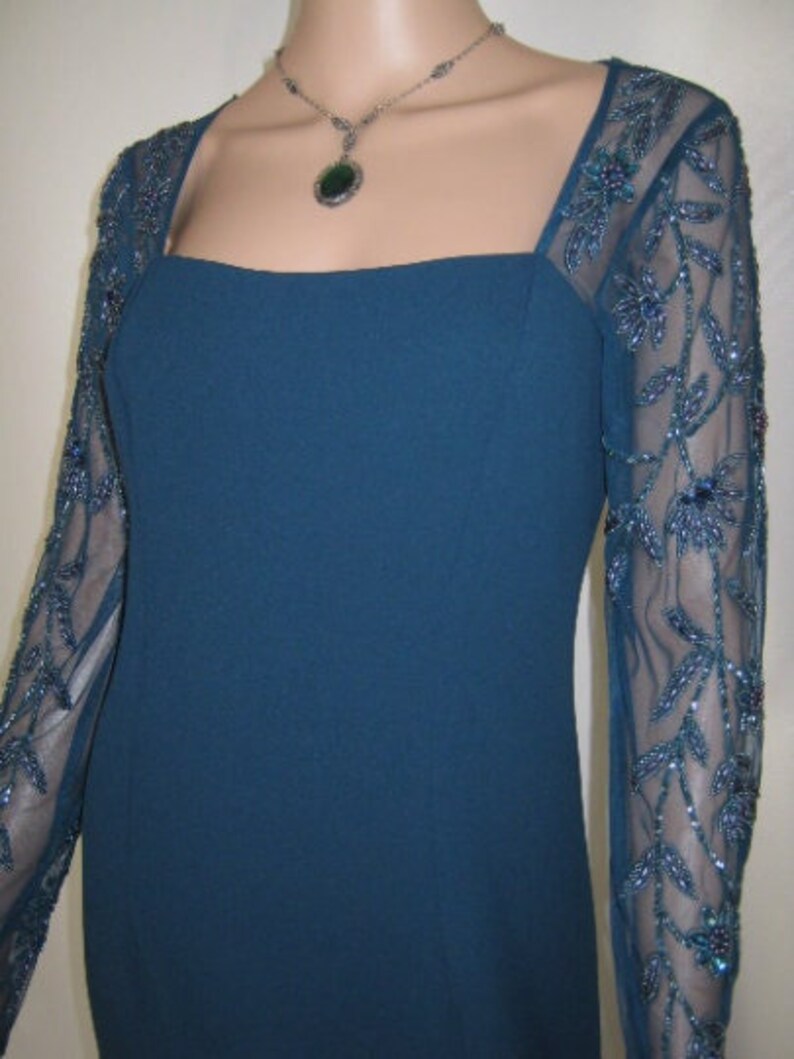 May include: A teal formal dress with a square neckline and sheer sleeves. The sleeves feature a floral beaded design. A silver necklace with a green pendant complements the dress.