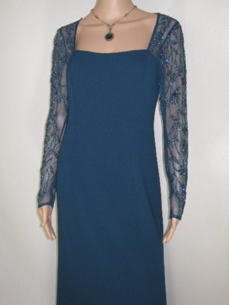 May include: A teal formal gown with a square neckline and sheer sleeves embellished with blue beading. The floor-length dress is accessorized with a silver necklace and green pendant. The dress is displayed on a mannequin.