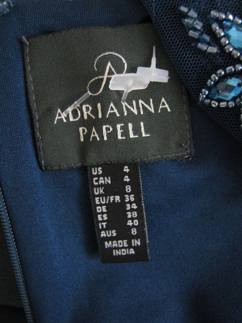 May include: A teal dress label with the brand name "ADRIANNA PAPELL" in white. The label shows size conversions for US, CAN, UK, EU/FR, DE, ES, IT, and AUS. The dress is made in India.