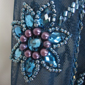 May include: Close-up of a dark blue mesh fabric with a floral embellishment. The flower has large, faceted blue gems, small silver beads, and clusters of purple pearls. Additional blue beadwork enhances the design.