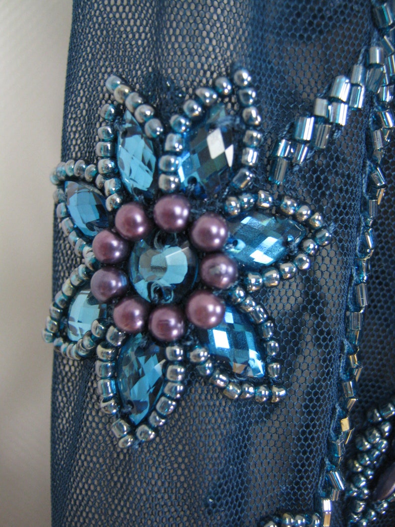May include: Close-up of a dark blue mesh fabric with a floral embellishment. The flower has large, faceted blue gems, small silver beads, and clusters of purple pearls. Additional blue beadwork enhances the design.