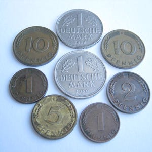 Può includere: Una collezione di monete tedesche vintage, tra cui monete da 1 e 10 Pfennig e 1 Deutsche Mark. Le monete sono in varie tonalità di bronzo e argento, con testo e date visibili.