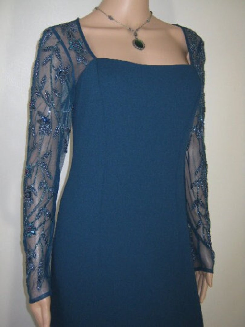 May include: A teal formal dress with a square neckline and long, sheer sleeves. The sleeves feature detailed beadwork. A silver necklace with a pendant is also visible.