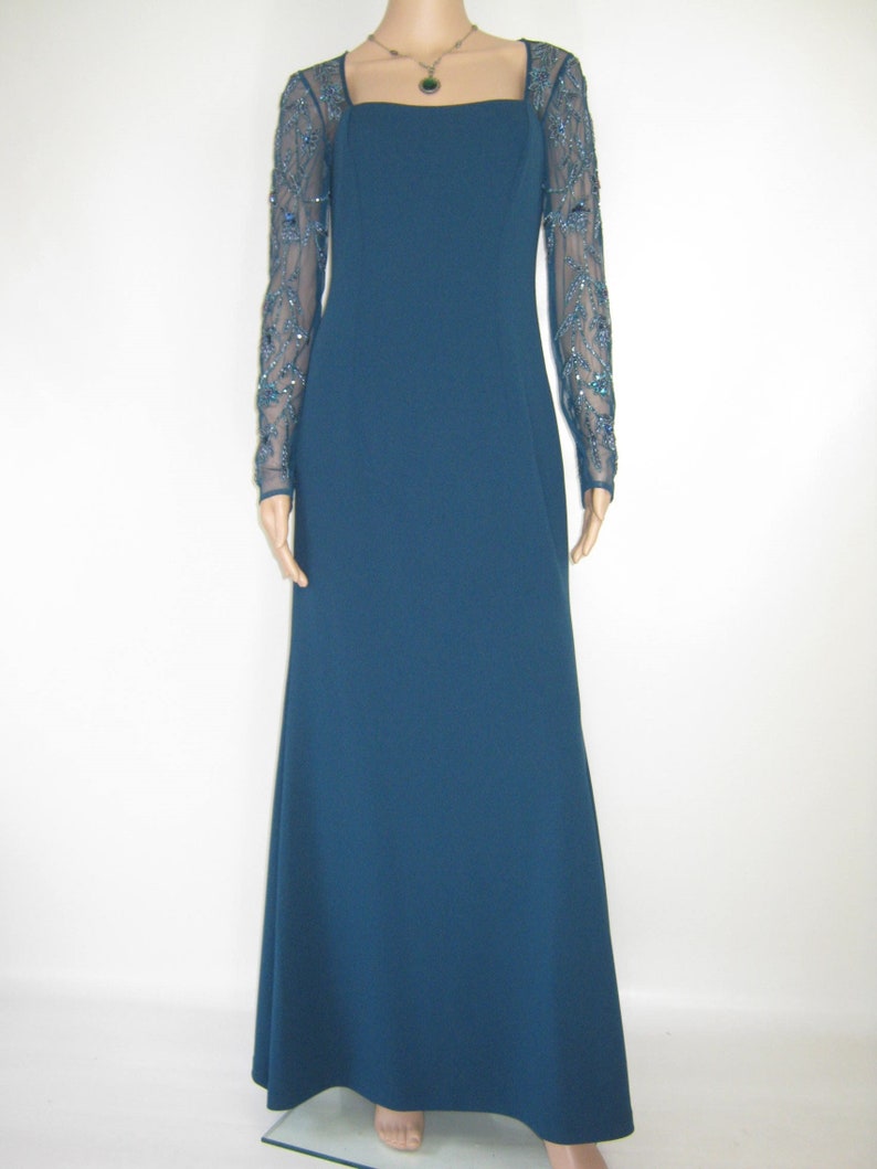 May include: A teal evening gown with a square neckline and long sleeves. The sheer sleeves are embellished with detailed beadwork. A delicate necklace with a pendant complements the dress. The dress has a floor-length skirt.