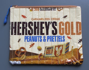 May include: A beige zippered pouch made from recycled Hershey's Gold chocolate wrappers. The pouch features the Hershey's Gold logo, the text "Caramelized Creme, Peanuts & Pretzels", and an image of a pretzel and peanuts.