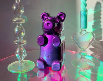 May include: Three iridescent glass candle holders with a purple gummy bear in the centre. The candle holders are in the shape of a heart and a tall cylinder. The gummy bear is sitting on a reflective surface.