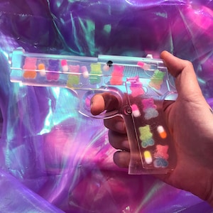 May include: A clear plastic toy gun filled with colorful gummy bears. The gun is held in a hand against a purple iridescent background.