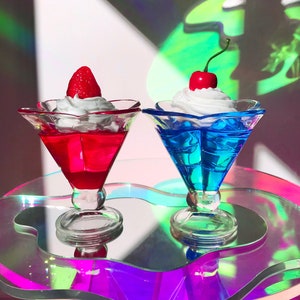 K&ouml;nnte beinhalten: Zwei klare Glasdessertschalen mit sternf&ouml;rmigem Rand, gef&uuml;llt mit roter und blauer Gelee, gekr&ouml;nt mit Schlagsahne und einer Kirsche und einer Erdbeere. Die Schalen stehen auf einem schillernden, wellenf&ouml;rmigen, klaren Glastisch.