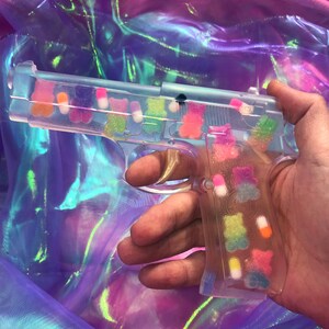 May include: A clear plastic toy gun filled with colorful gummy candies. The gun is held in a hand against a iridescent background.