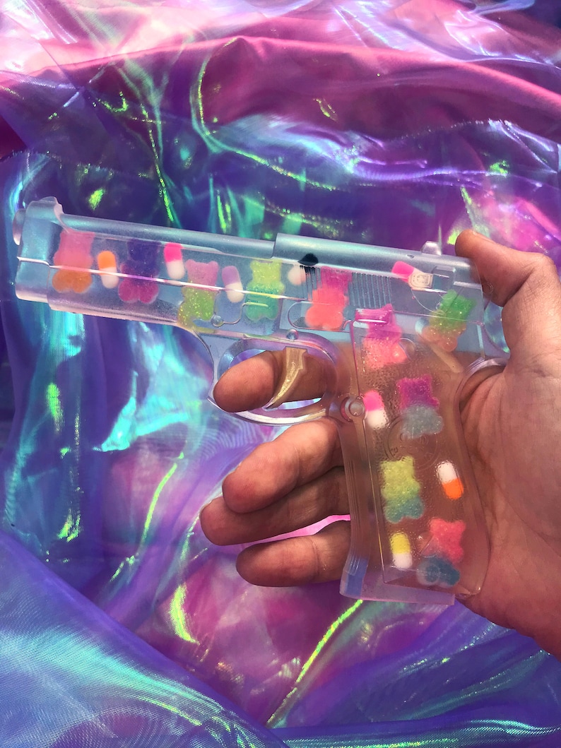 May include: A clear plastic toy gun filled with colorful gummy candies. The gun is held in a hand against a iridescent background.
