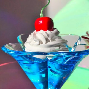 K&ouml;nnte beinhalten: Eine blaue Glassch&uuml;ssel mit einem Topping aus wei&szlig;er Schlagsahne und einer roten Kirsche.
