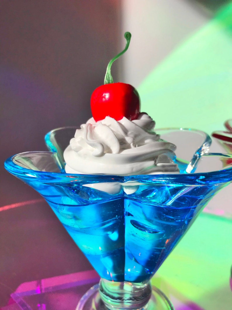 K&ouml;nnte beinhalten: Eine blaue Glassch&uuml;ssel mit einem Topping aus wei&szlig;er Schlagsahne und einer roten Kirsche.