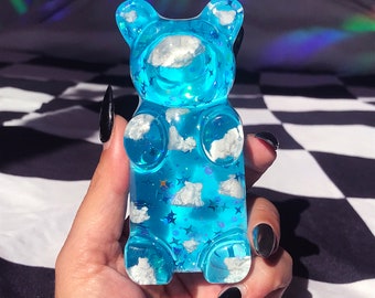 May include: A translucent blue gummy bear figurine filled with white cloud shapes and blue star-shaped glitter. The bear is held up against a black and white checkered background, with rainbow light reflections in the background.