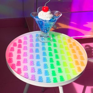 May include: A white side table with three legs, featuring a rainbow gradient of gummy bears on the tabletop. A glass dessert dish with a blue liquid and whipped cream topped with a red cherry sits on the table.