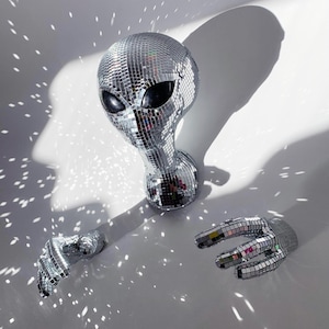 Puede incluir: Una cabeza de extraterrestre de bola de discoteca plateada con azulejos de espejo. El extraterrestre tiene ojos grandes y una cabeza redonda. El extraterrestre está sobre un fondo blanco con luz reflejándose en los azulejos de espejo.