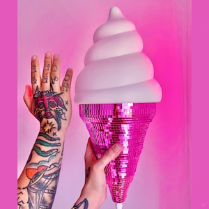 May include: A whimsical ice cream cone lamp with a white, swirled top and a pink, mirrored cone base. The lamp is held against a pink background. The lamp has a white cord.
