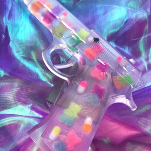 May include: A clear plastic toy gun filled with colorful candy-like objects. The gun is lying on a iridescent purple and blue fabric.