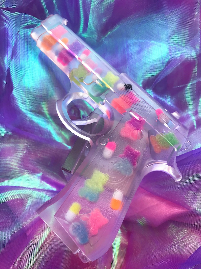 May include: A clear plastic toy gun filled with colorful candy-like objects. The gun is lying on a iridescent purple and blue fabric.