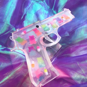 May include: A clear plastic gun-shaped mold filled with colorful gummy candies. The mold is on a purple iridescent fabric background.