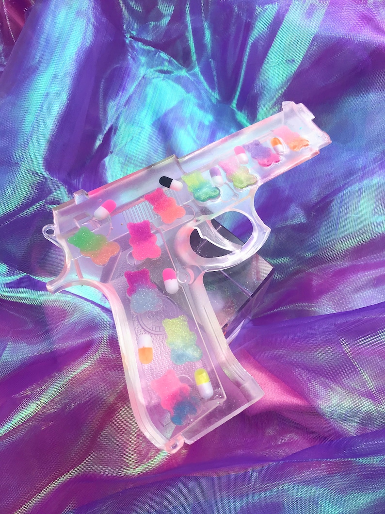 May include: A clear plastic gun-shaped mold filled with colorful gummy candies. The mold is on a purple iridescent fabric background.