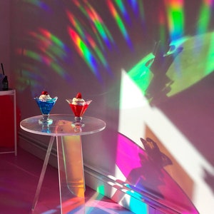K&ouml;nnte beinhalten: Zwei klare Glasschalen mit Schlagsahne und Fruchtbelag stehen auf einem klaren Acryltisch mit regenbogenfarbenem, irisierendem Finish. Der Tisch hat drei Beine und steht auf einer rosa Oberfl&auml;che. Der Hintergrund ist eine wei&szlig;e Wand mit Regenbogenlichtreflexionen.