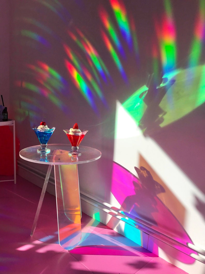 K&ouml;nnte beinhalten: Zwei klare Glasschalen mit Schlagsahne und Fruchtbelag stehen auf einem klaren Acryltisch mit regenbogenfarbenem, irisierendem Finish. Der Tisch hat drei Beine und steht auf einer rosa Oberfl&auml;che. Der Hintergrund ist eine wei&szlig;e Wand mit Regenbogenlichtreflexionen.