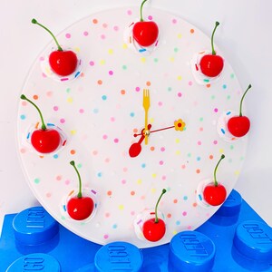 May include: A white clock with a colorful confetti design and red cherry accents. The clock is sitting on top of blue LEGO blocks.