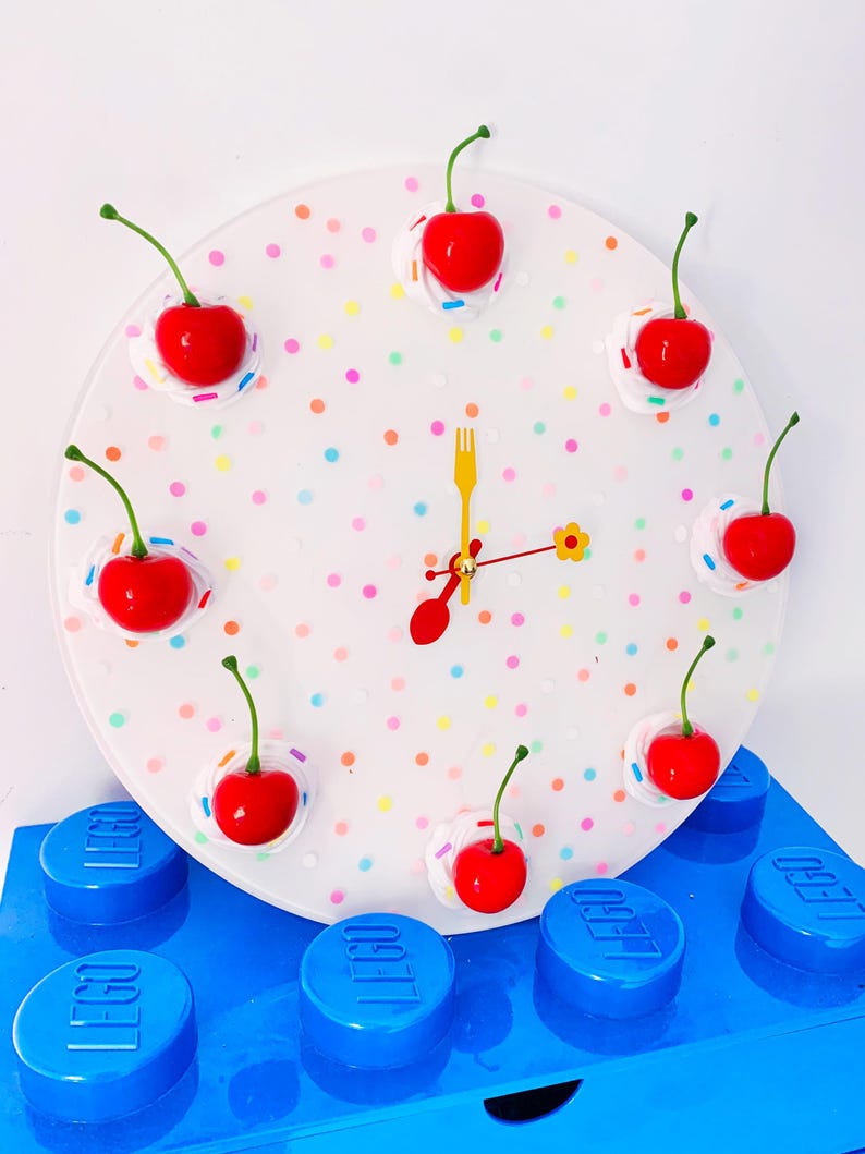 May include: A white clock with a colorful confetti design and red cherry accents. The clock is sitting on top of blue LEGO blocks.
