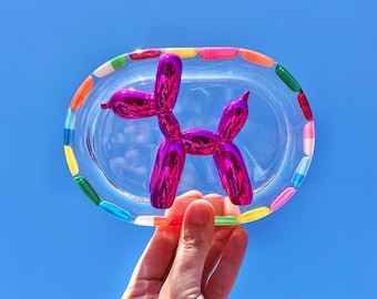 May include: A clear, oval-shaped tray with a colorful border of rainbow-colored segments. A shiny, metallic magenta balloon dog sculpture sits in the center. The tray is held up against a bright blue sky.