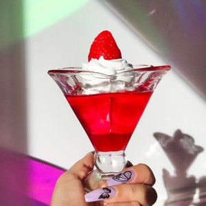 K&ouml;nnte beinhalten: Eine klare Glassch&uuml;ssel gef&uuml;llt mit roter Gelee, die mit Schlagsahne und einer roten Erdbeere garniert ist. Die Sch&uuml;ssel wird von einer Hand mit lila Nagellack und schwarzen Schmetterlings-Nagelkunst gehalten.