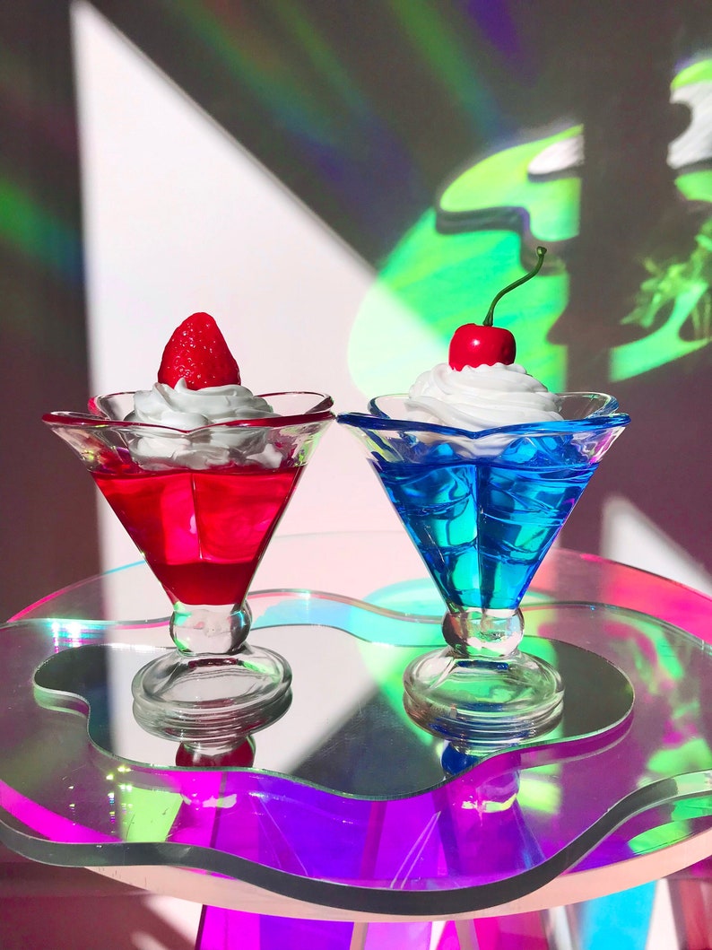 K&ouml;nnte beinhalten: Zwei klare Glasdessertschalen mit sternf&ouml;rmigem Rand, gef&uuml;llt mit roter und blauer Gelee, gekr&ouml;nt mit Schlagsahne und einer Kirsche und einer Erdbeere. Die Schalen stehen auf einem schillernden, wellenf&ouml;rmigen, klaren Glastisch.