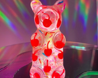 May include: A clear gummy bear figurine with red cherries and sprinkles inside. The bear is standing on a reflective surface.