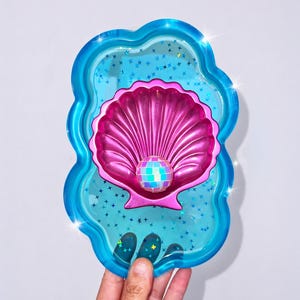 May include: A decorative tray with a scalloped edge, featuring a pink seashell design with a disco ball center. The tray is filled with blue glitter and star-shaped confetti, and has a bright blue border.