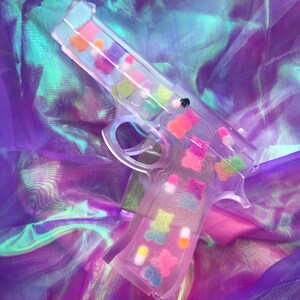 May include: A clear plastic toy gun filled with colorful gummy bears and candy. The gun is lying on a iridescent purple and blue fabric.
